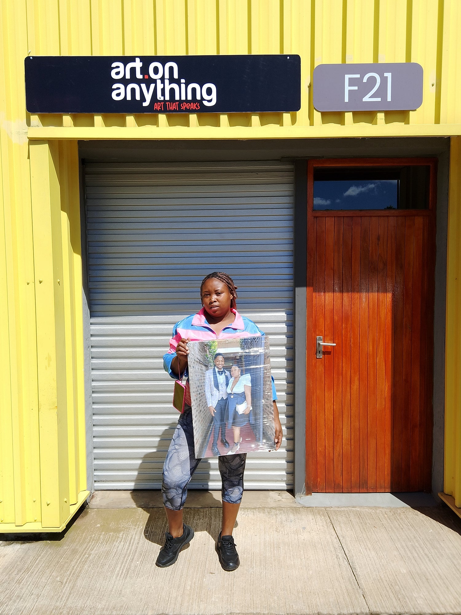 Person holding a framed picture in front of a yellow building with 'art on anything' signage.